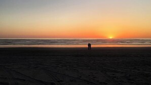 A beach with footprints leading to the water, a sunset in the background, and two people standing at the water's edge.