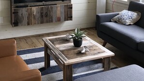 A living room with a television mounted on a rustic wooden wall, a sofa with a cushion, a coffee table with decorative items, and a striped rug.