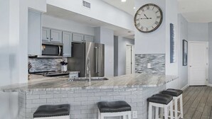 A modern kitchen with a marble island, stainless steel appliances, and a large wall clock. - Guest Favorite| Gulf Front | Accommodates 12 |  Beach Serv. I Pet Friendly| (Miramar Beach)