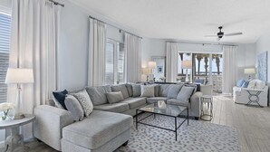 A modern living room with a sectional sofa, a glass coffee table, and a view of palm trees through large windows. - Guest Favorite| Gulf Front | Accommodates 12 |  Beach Serv. I Pet Friendly| (Miramar Beach)