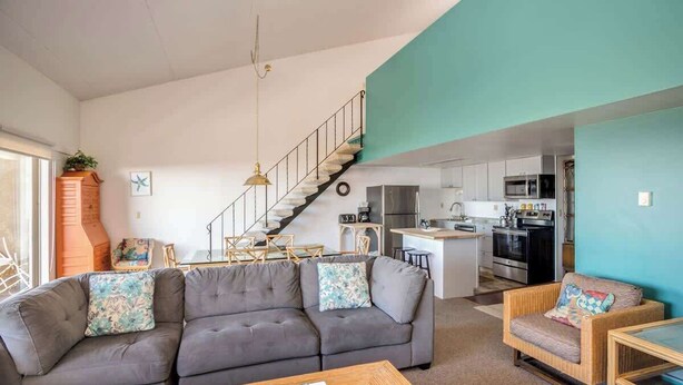 A living room with a grey sofa, a wooden coffee table, a wicker armchair, a staircase, and a kitchen in the background. - Gearhart House G748 (Gearhart)