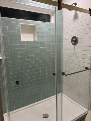 A glass-enclosed shower with a tiled wall, a shower control panel, and a handrail.