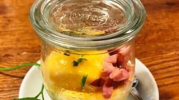 Un frasco de vidrio con tapa que contiene una preparación en capas, posiblemente una ensalada o aperitivo, con ingredientes visibles como huevos, jamón y hierbas, colocado sobre un plato blanco con una cuchara.