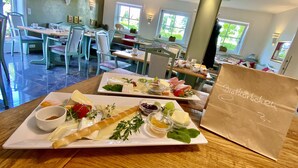 A dining area with tables set for a meal, featuring a variety of cheeses, fruits, and a breadstick. A paper bag with the name 'Brothöber' is visible. - Hotel Auszeit (Isernhagen)