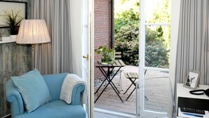 A room with a blue sofa, a lamp, a desk with a phone and papers, and a view of an outdoor patio with a table and chairs. - Hotel Auszeit (Isernhagen)