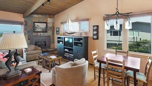 Un salon avec une cheminée, une table à manger avec des chaises, un canapé, un fauteuil en osier et une lampe.