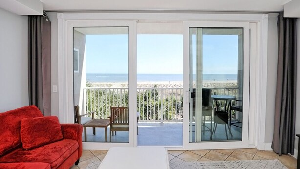 A room with a red sofa, a white coffee table, and a view of the beach through glass doors. - Beachfront Condo 322, Luxury 1 BR at Beachside Colony NEW POOL (Tybee Island)
