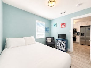 A bedroom with a bed, a TV, a chair, and a kitchen visible through an open door.