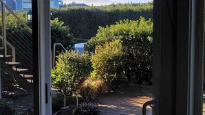 A view through a glass door of a garden area with a wooden deck, a staircase, and a hedge.