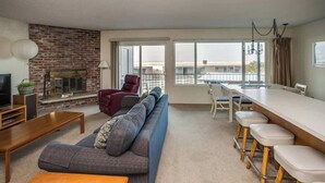 A modern living room with a brick fireplace, a large sofa, a dining area with bar stools, and large windows offering a view. - Pacific View V473 (Gearhart)