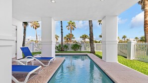 A swimming pool with blue and white lounge chairs, surrounded by palm trees and a white fence. - Incredible Beach House with Pool! Short safe walk to the beach, Family Area (South Padre Island)