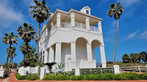 A white colonial-style building with a balcony, surrounded by palm trees and a red pathway. - Incredible Beach House with Pool! Short safe walk to the beach, Family Area (South Padre Island)
