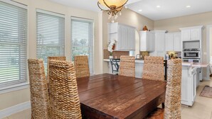 A dining room with a wooden table and woven chairs, a kitchen with white cabinets, and a chandelier. - Incredible Beach House with Pool! Short safe walk to the beach, Family Area (South Padre Island)