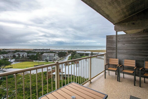 Un balcón con vista a la playa y casas, sillas de madera y una mesa.