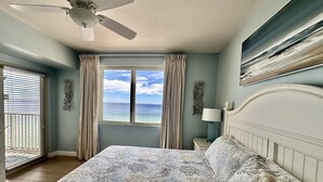 A bedroom with a large bed, a ceiling fan, a window with a view of the ocean, and a painting on the wall.