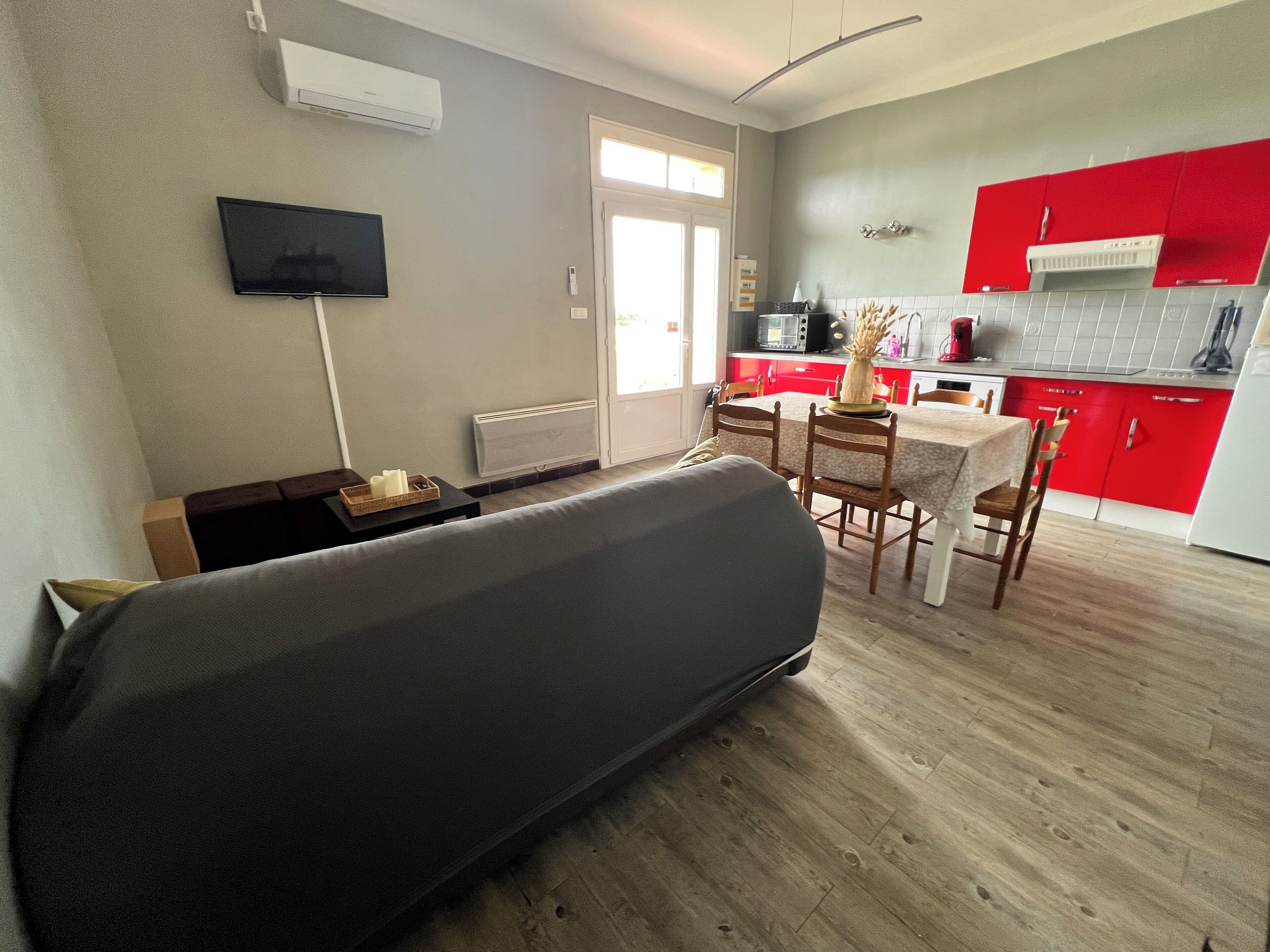 A living room with a sofa, a TV mounted on the wall, a dining area with a table and chairs, and a kitchen with red cabinets and a white refrigerator.