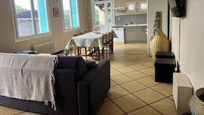 A living room with a sofa, a dining table with chairs, a kitchen area with cabinets and a refrigerator, and large windows with blinds.