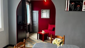 A room with a checkered floor, a dining table with chairs, a vase with flowers, a red sofa, and a door leading to a kitchen.