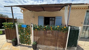 A covered outdoor area with a fence, potted plants, and a building with a window and shutters.
