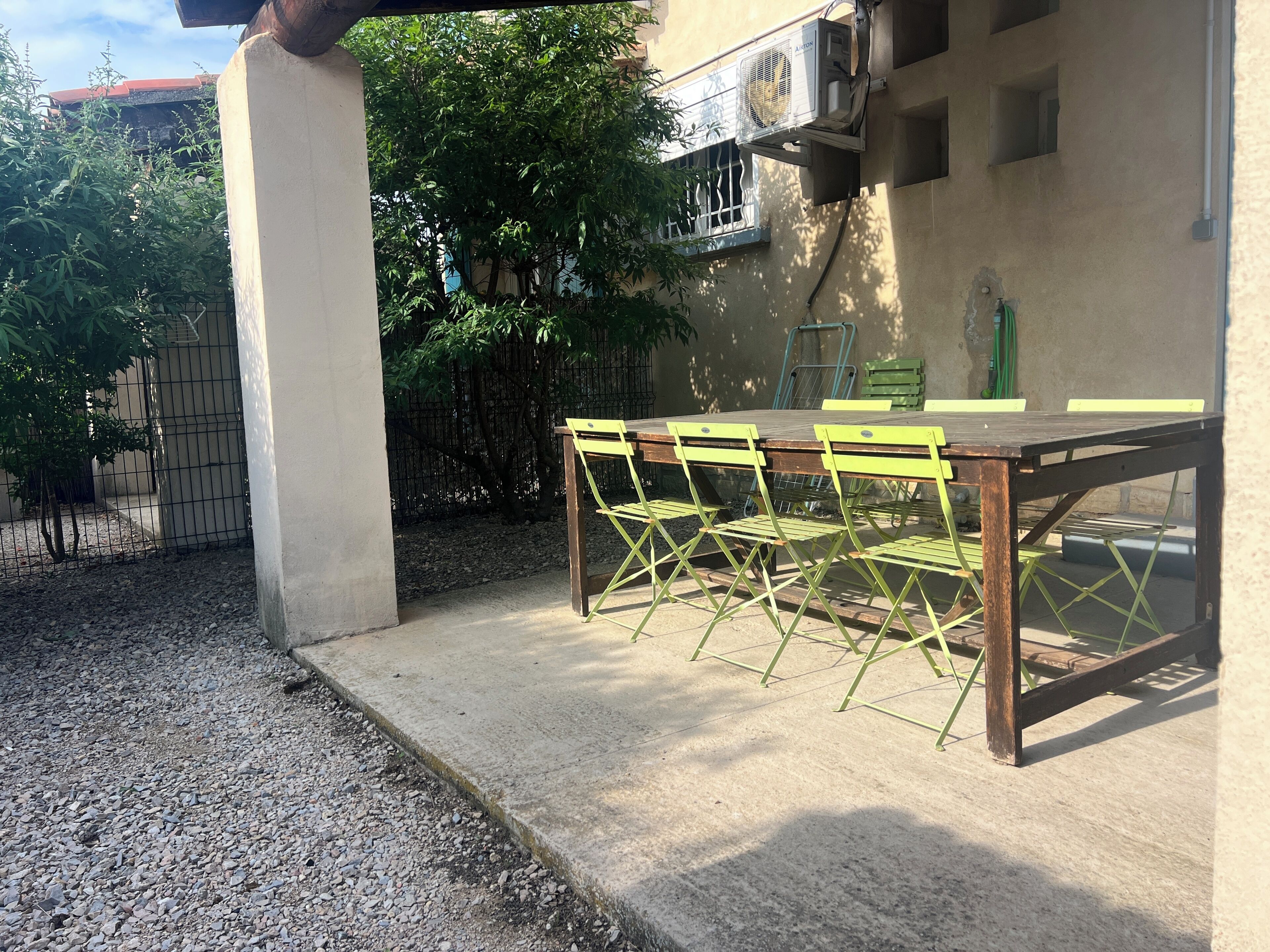 En udendørs terrasse med et betonbord og grønne stole, en vægmonteret aircondition og en metalport.