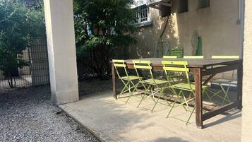 En udendørs terrasse med et betonbord og grønne stole, en vægmonteret aircondition og en metalport.