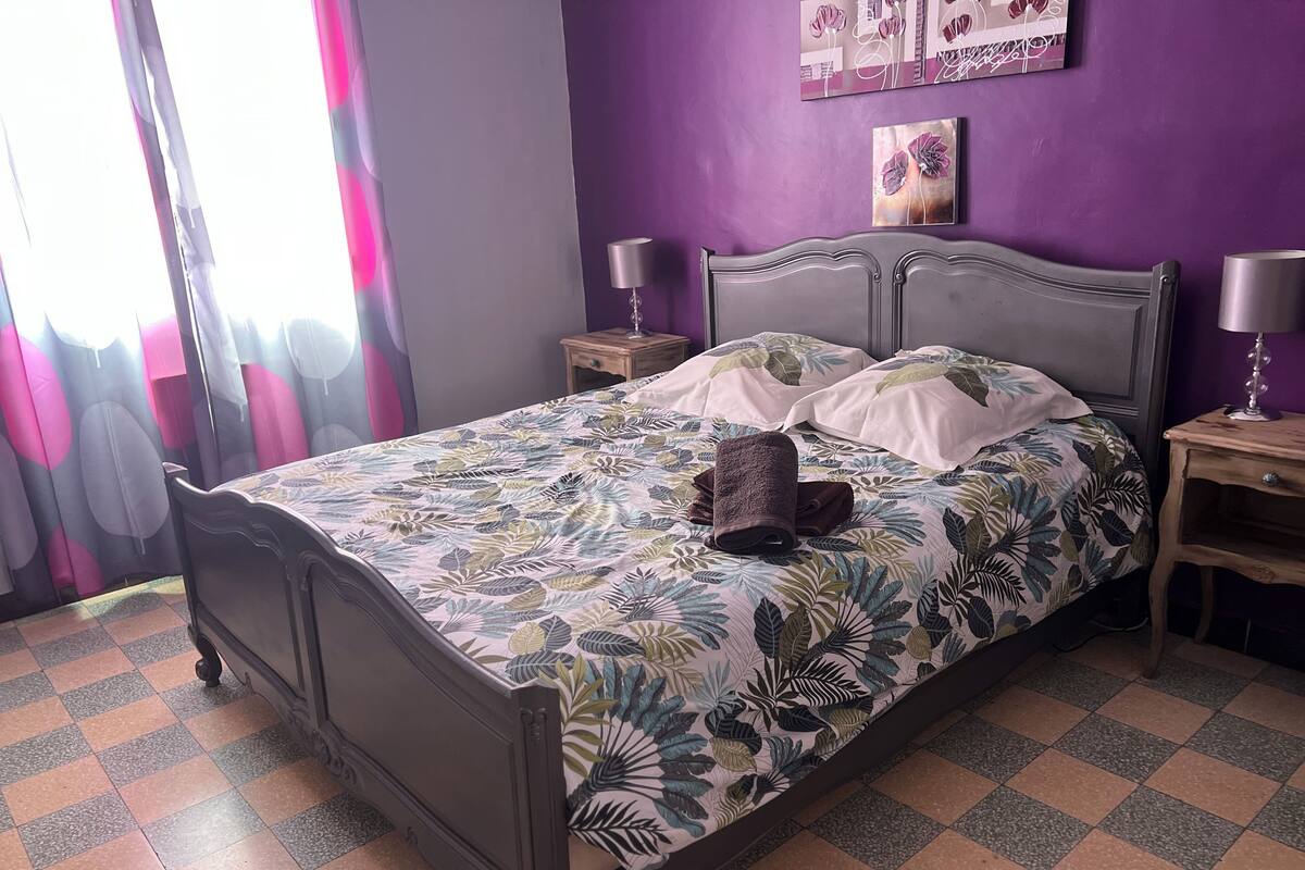 Una cama con colcha estampada, dos mesitas de noche con lámparas, una ventana con cortinas y una pared decorada con obras de arte.