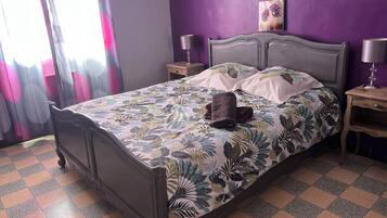 Un lit recouvert d’une couvre-lit à motifs floraux, deux tables de chevet avec des lampes, une fenêtre avec des rideaux et un mur orné d’œuvres d’art.
