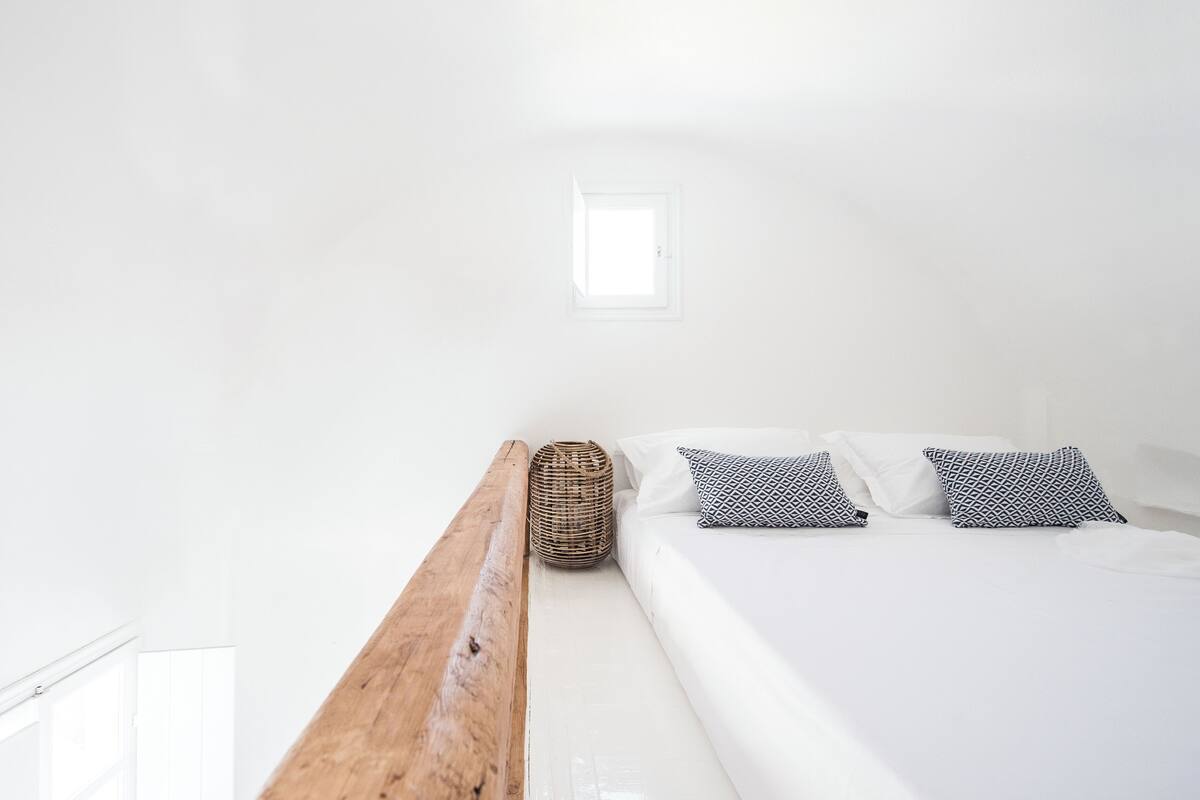 Un dormitorio minimalista con cabecera de madera, cama con ropa blanca y almohadas estampadas, y una ventana pequeña.