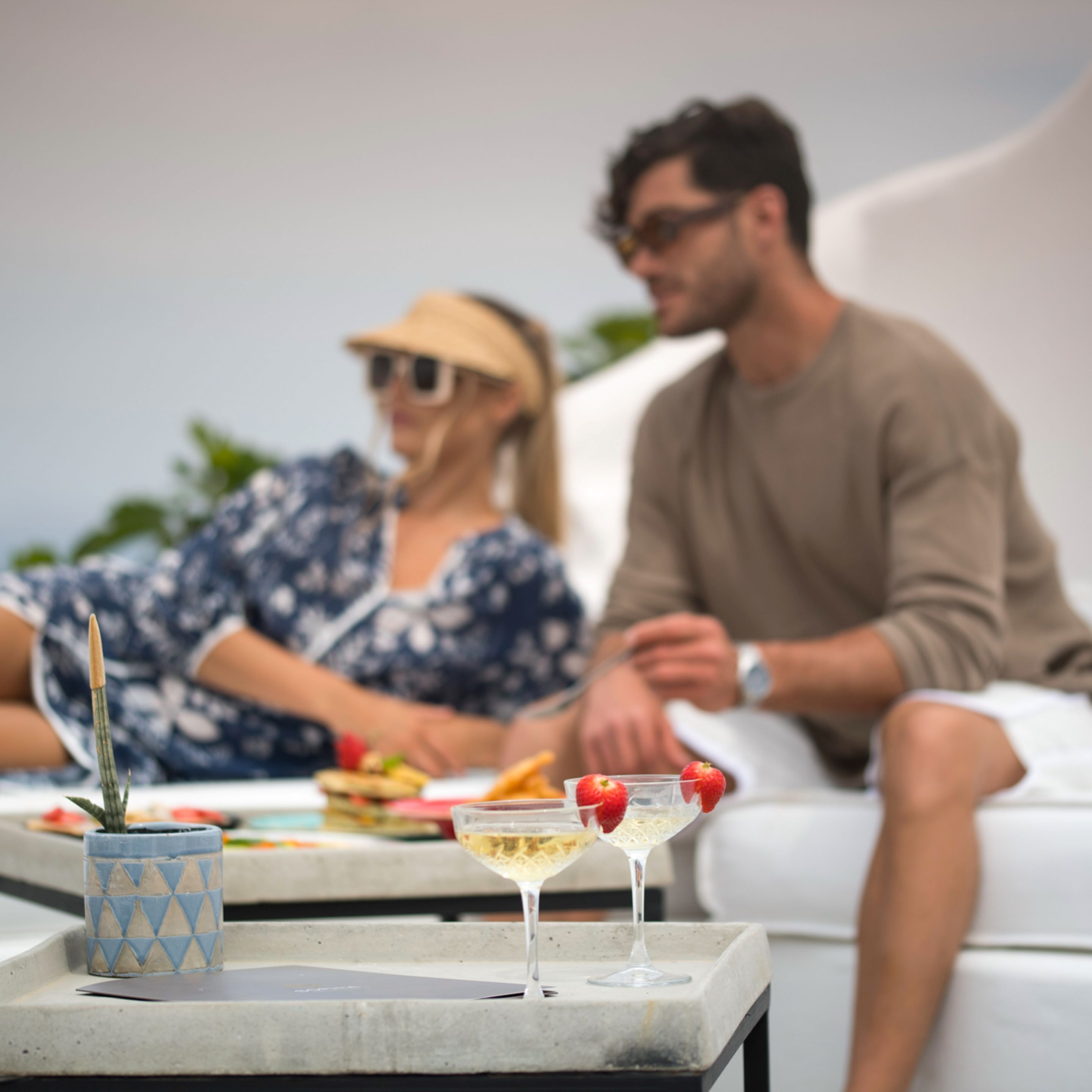 Una pareja sentada en un sofá blanco al aire libre, con una mesa frente a ellos donde hay bebidas y una pequeña planta.