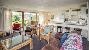 Ein Wohnzimmer mit Glastisch, Korbstühlen, einem braunen Sofa und einer Küchenzeile mit weißen Schränken.
