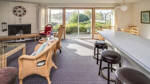 Ein modernes Wohnzimmer mit Kamin, Korbmöbeln, einem Essbereich und einem großen Schiebeglasfenster, das in einen Garten führt.