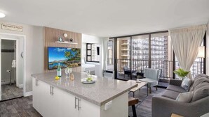 A modern living room with a kitchen island, a dining area, and large windows with a city view. - Private lanai & heated pool - 1 block to the beach (Honolulu)