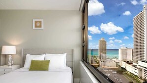 A hotel room with a bed, a nightstand, a lamp, and a window with a city view. - Private lanai & heated pool - 1 block to the beach (Honolulu)