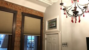 Ein Zimmer mit Backsteinwand, zwei Fenstern mit Jalousien, einer weißen Tür, einer Kandelaber und einem kleinen Buntglasfenster über der Tür.