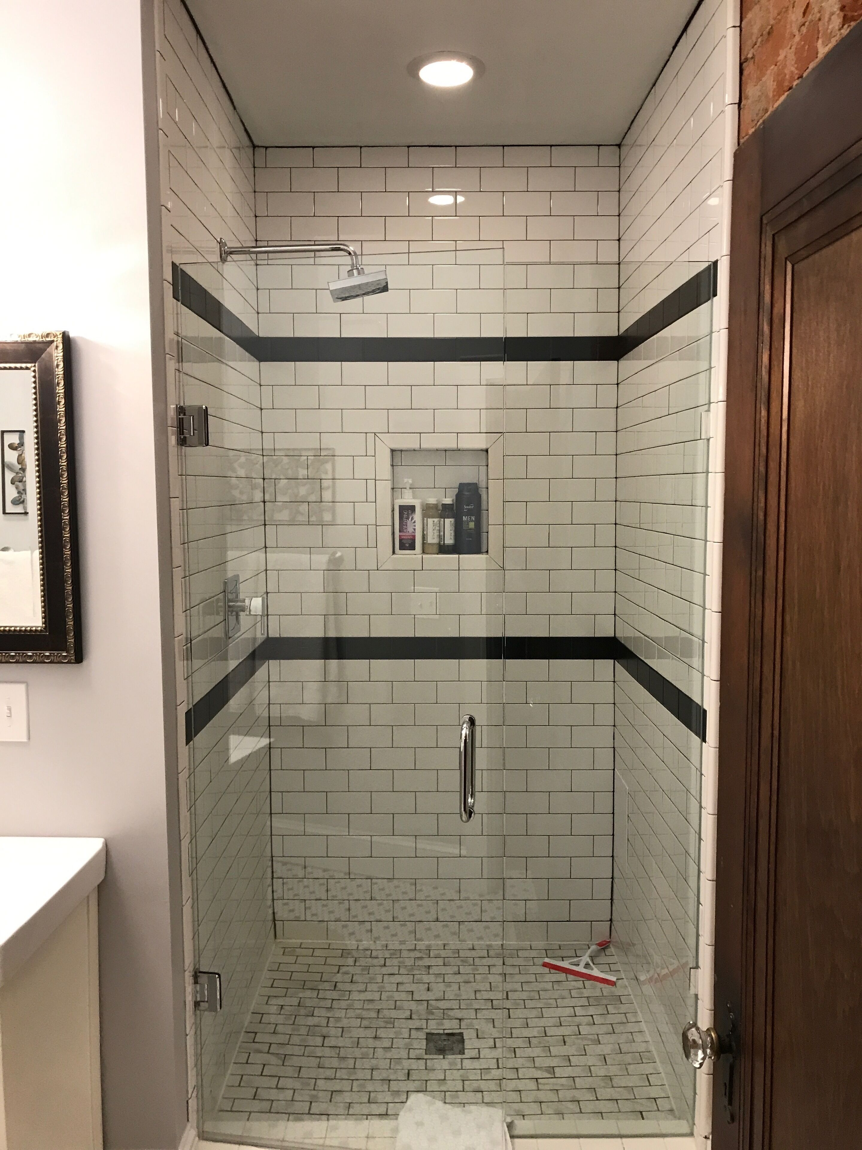 A shower with white and black tiles, a glass door, and a small shelf with toiletries.