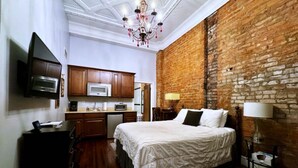Ein Schlafzimmer mit Backsteinwand, einem Bett mit weißer Bettwäsche, einer Kristallleuchtre, einem Flachbildfernseher und einer Küchenzeile.