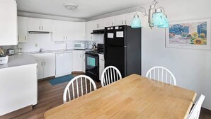 A compact kitchen with white cabinets, a black refrigerator, a wooden dining table, and white chairs. - Pacific Terrace T443 (Gearhart)