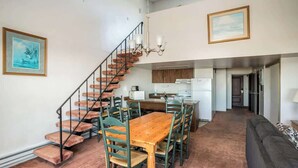Een ruime woonkamer met een houten eettafel, groene stoelen, een trap en een keuken op de achtergrond.