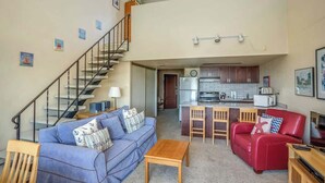 A two-story apartment with a staircase, a living room with a blue sofa and red leather chair, a kitchen area, and a small dining table. - Gearhart House G700 (Gearhart)