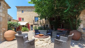 An outdoor seating area with wicker furniture and a central fire pit, surrounded by stone buildings and greenery. - La Louvière des Bruyères (Robion)