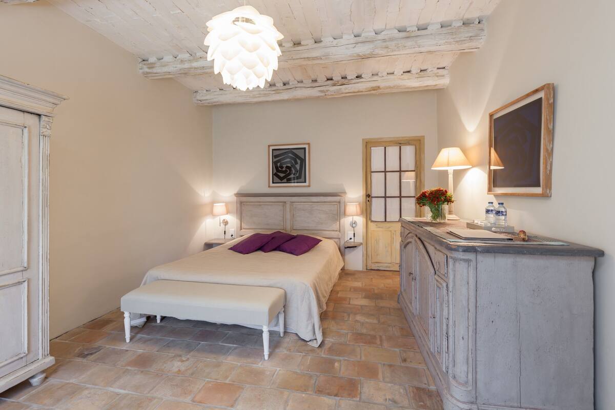 A bedroom with a bed, bench, dresser, and a hanging light fixture.