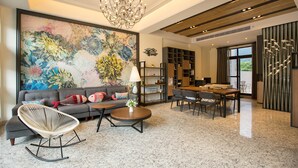 Uma sala de estar ampla com um grande quadro floral, um sofá moderno, uma poltrona de balanço única, uma mesa de jantar com cadeiras e um lustre.
