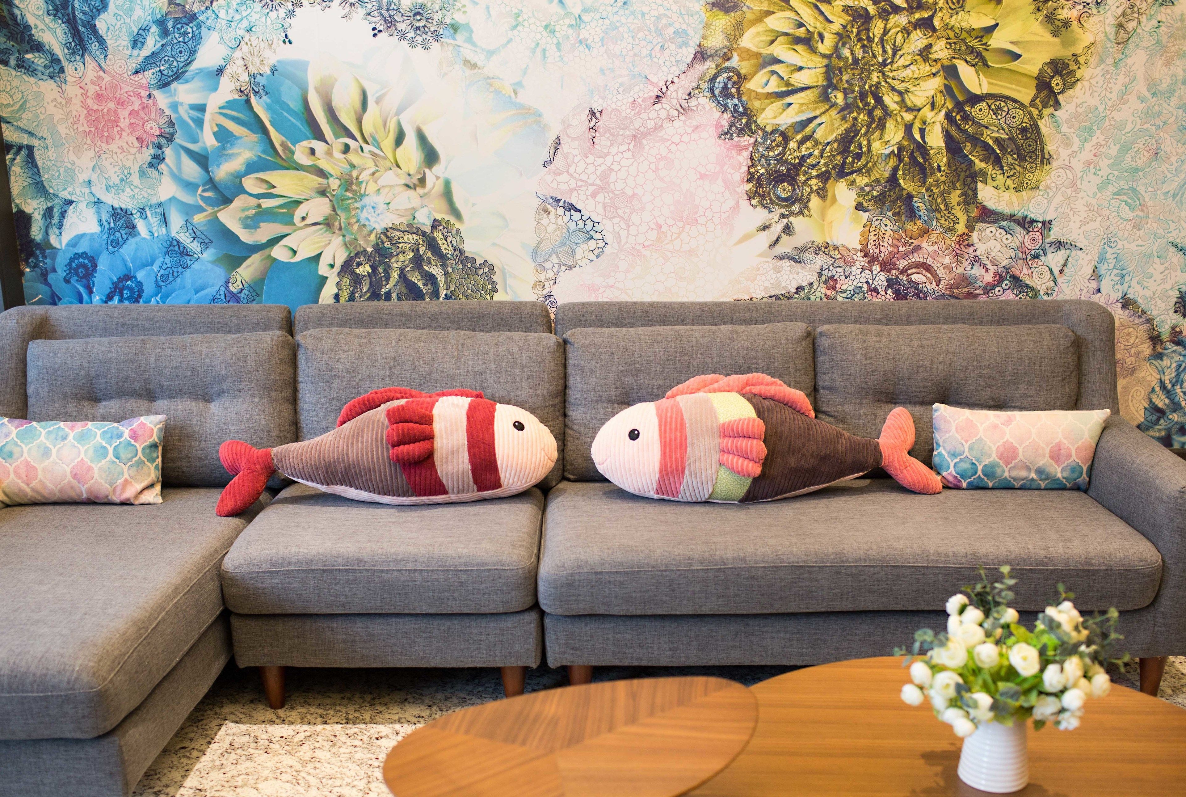 A grey sofa with two fish-shaped pillows and a white vase with flowers on a wooden table.