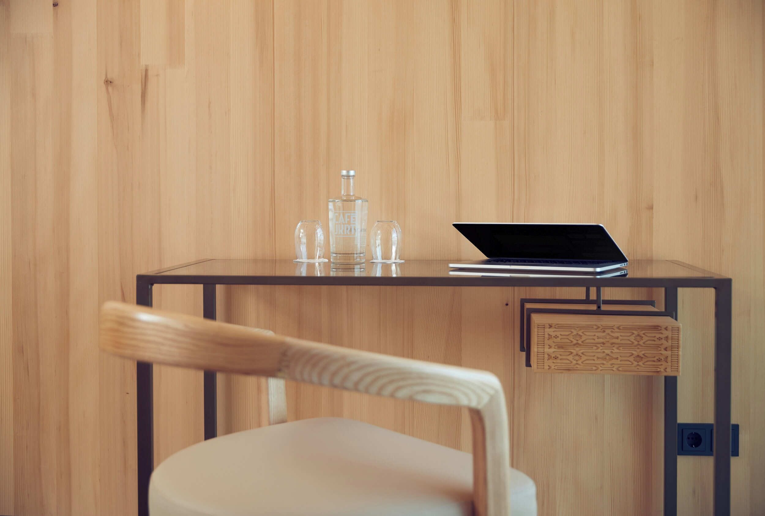 A wooden wall, a glass bottle, two glasses, a laptop, and a chair with a wooden backrest.