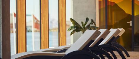 A row of sun loungers with curved black legs, positioned in front of a large window with a view of a mountain range.