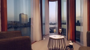 Habitación de hotel con vista a un horizonte urbano y una masa de agua. Hay una mesa redonda con una botella de champán y una copa.