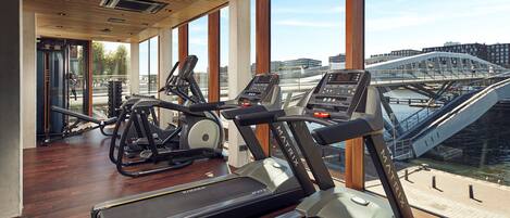 A gym with treadmills and elliptical machines, large windows, and a view of a bridge and buildings.