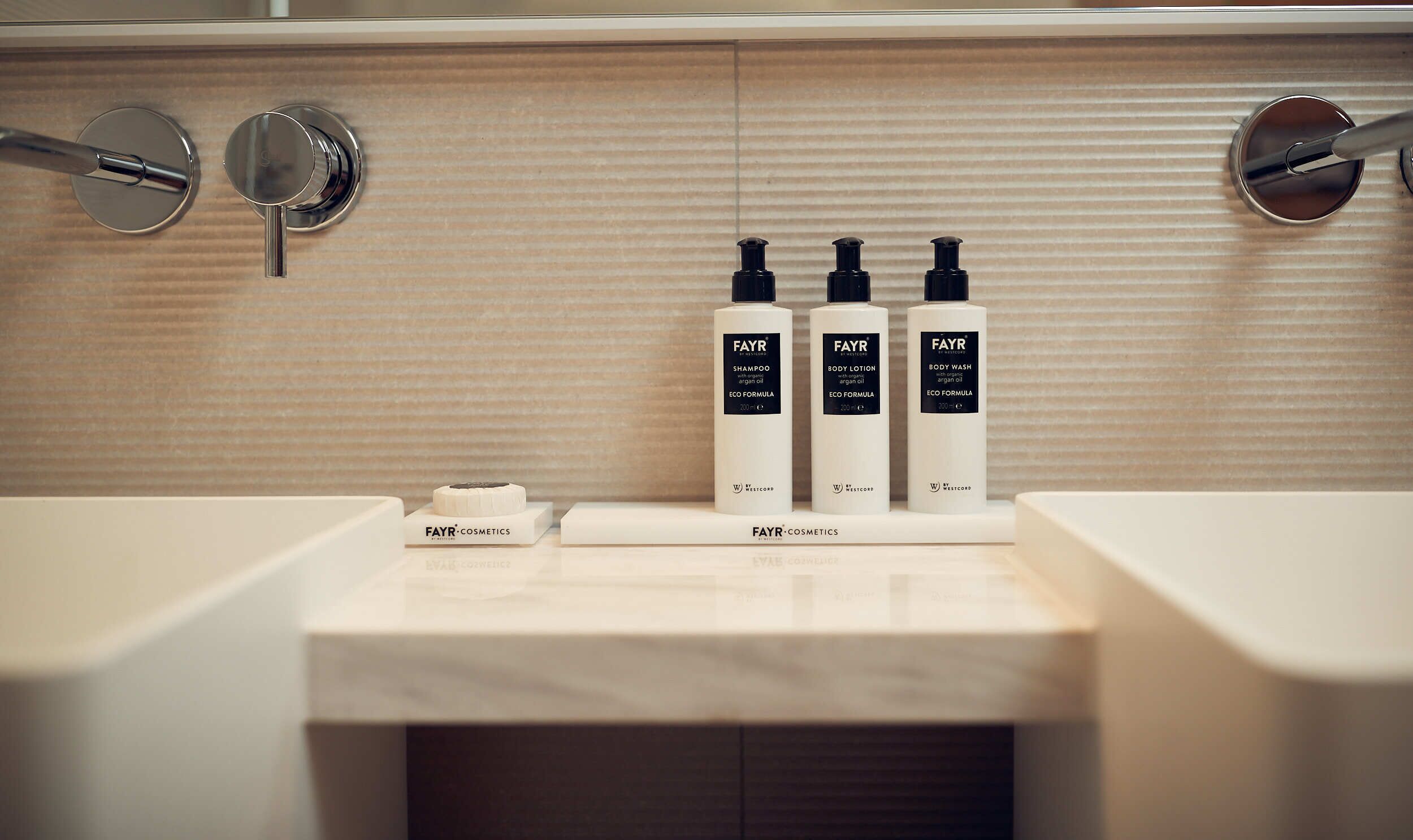 Une salle de bain avec un comptoir en marbre, trois flacons de produits de la marque FAYR Cosmetics et un porte-savon.