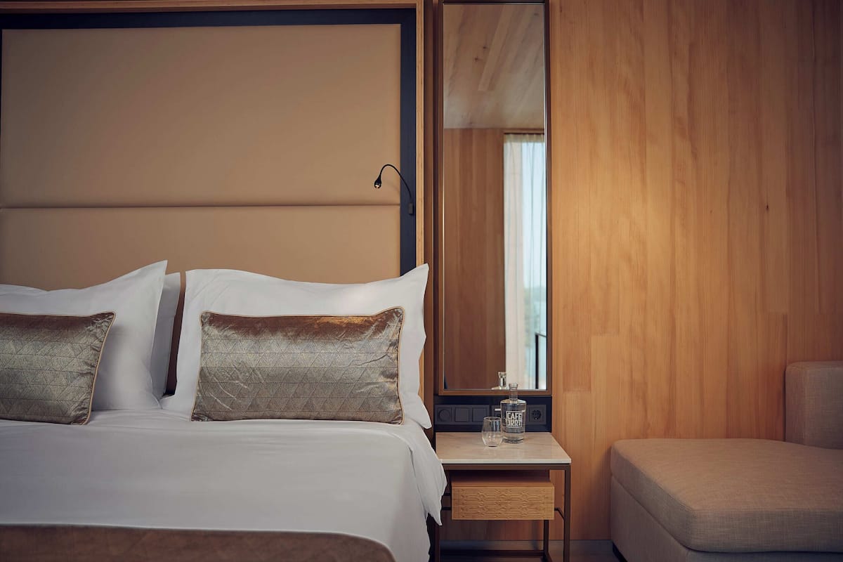 Un lit bien fait, avec du linge de lit blanc et deux coussins décoratifs, une table de chevet avec une bouteille et un verre, et une table de nuit en bois.