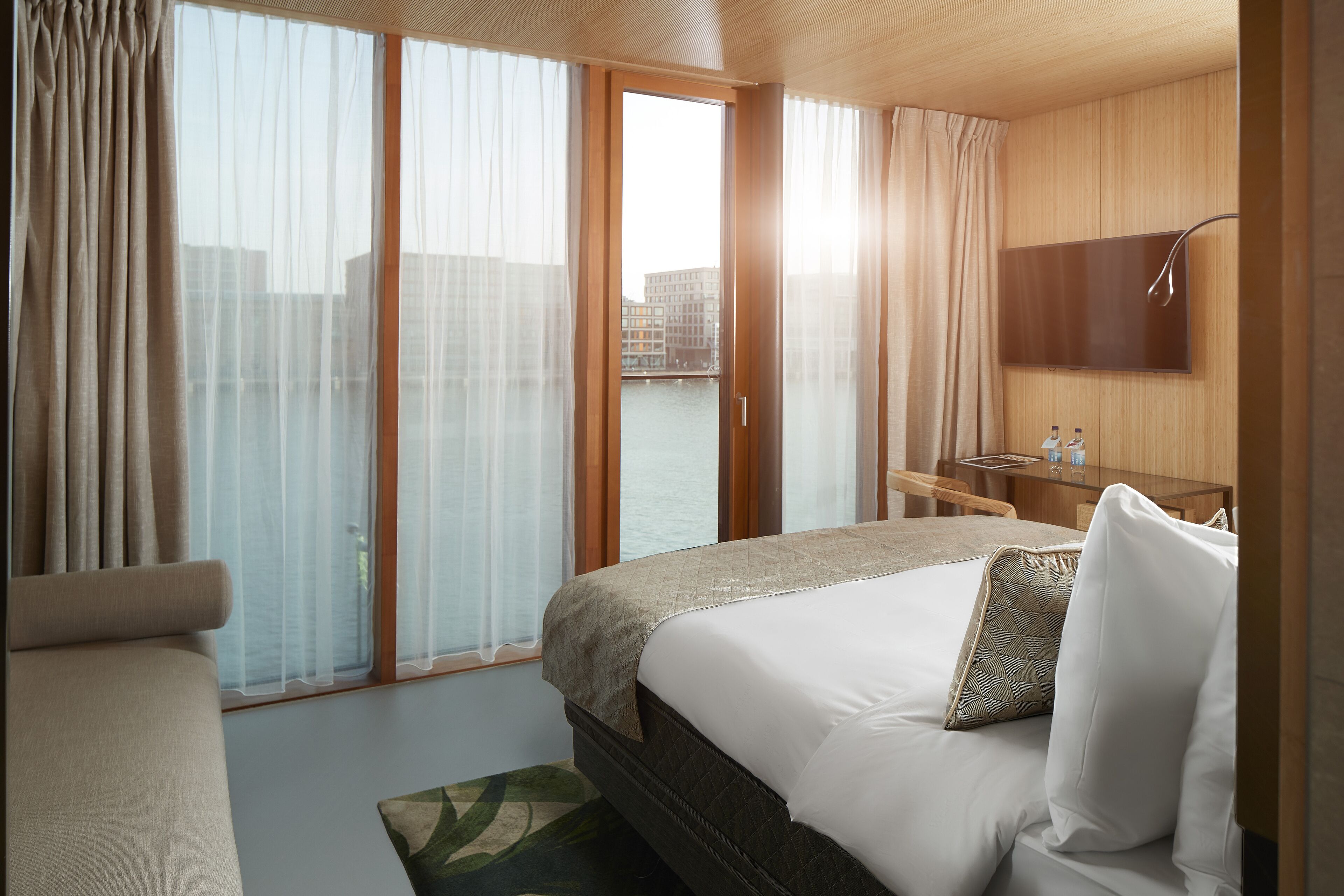 Habitación de hotel con una cama grande, un sofá, un televisor montado en la pared y vistas al agua a través de la ventana.